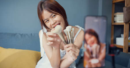 Happy young Asian girl makeup vlogger front of phone camera enjoy review talk with audience in living room at house. Social distance coronavirus pandemic concept. Freedom and active lifestyle concept.の写真素材
