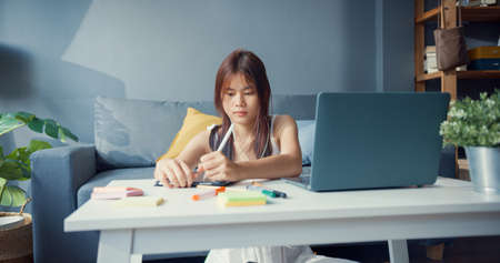 Young Asia girl teenager with casual shirt wear earphone use laptop learn online write lecture in laptop in living room at house. Isolate education online e-learning coronavirus pandemic concept.の写真素材