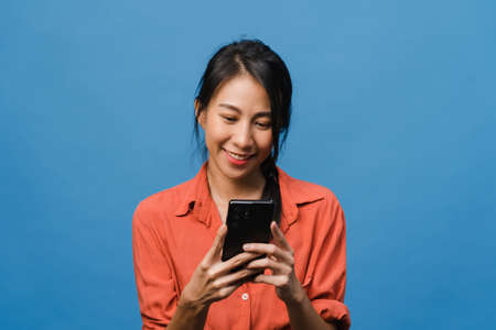 Young Asia lady using phone with positive expression, smiles broadly, dressed in casual clothing feeling happiness and stand isolated on blue background. Happy adorable glad woman rejoices success.の写真素材
