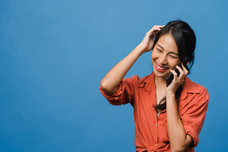 Young Asia lady talk by phone with positive expression, smile broadly, dressed in casual clothing feeling happiness and stand isolated on blue background. Happy adorable glad woman rejoices success.の写真素材