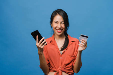 Young Asia lady using phone and credit bank card with positive expression, smiles broadly, dressed in casual clothing and stand isolated on blue background. Happy adorable glad woman rejoices success.の写真素材