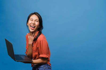 Young Asia lady using laptop with positive expression, smiles broadly, dressed in casual clothing feeling happiness and stand isolated on blue background. Happy adorable glad woman rejoices success.の写真素材