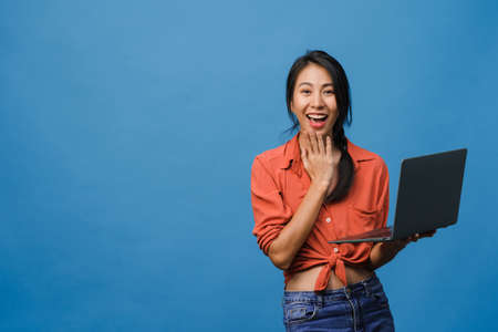 Young Asia lady show empty laptop screen with positive expression, smiles broadly, dressed in casual clothing feeling happiness isolated on blue background. Computer with white screen in female hand.の写真素材