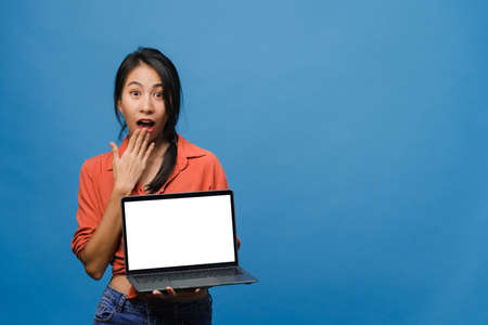 Young Asia lady show empty laptop screen with positive expression, smiles broadly, dressed in casual clothing feeling happiness isolated on blue background. Computer with white screen in female hand.の写真素材