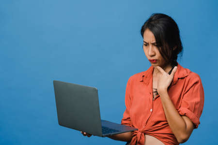 Young Asia lady using laptop with negative expression, excited screaming, cry emotional angry in casual cloth and stand isolated on blue background with blank copy space. Facial expression concept.の写真素材