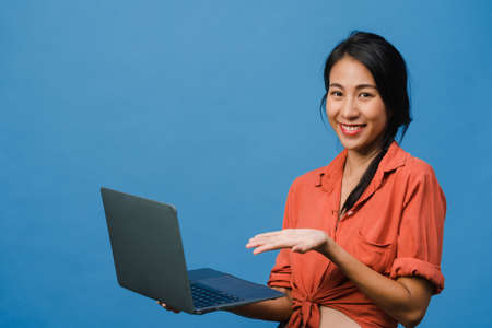 Young Asia lady using laptop with positive expression, smiles broadly, dressed in casual clothing feeling happiness and stand isolated on blue background. Happy adorable glad woman rejoices success.の写真素材