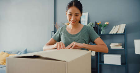 Happy beautiful Asia lady unboxing cardboard delivery package from online marketplace in living room at house. Satisfied shopper in internet unboxing goods, Online shopping and delivery concept.の写真素材