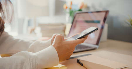 Close-up young asia lady freelance focus mobile phone chat with colleagues work finance graph account chart market plan in laptop at home. Girl student learn online at house, Work from home concept.の写真素材
