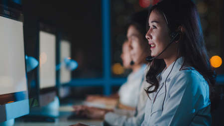 Millennial Asia young call center team or customer support service executive using computer and microphone headset working technical support in late night office. Telemarketing or sales job concept.の写真素材