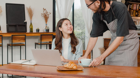 Young Asian freelance women work talk on phone with customer at coffee shop. Asian happy men barista waiter wear gray apon serve hot coffee to female customer who business work in cafe or restaurant.の写真素材