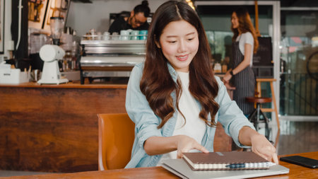 Cheerful Young Asian freelance lady working on laptop at coffee shop. Attractive Japanese businesswoman relax feeling happy in urban cafe or restaurant.の写真素材