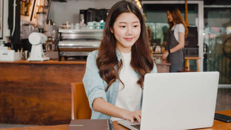 Cheerful Young Asian freelance lady working on laptop at coffee shop. Attractive Japanese businesswoman relax feeling happy in urban cafe or restaurant.の写真素材