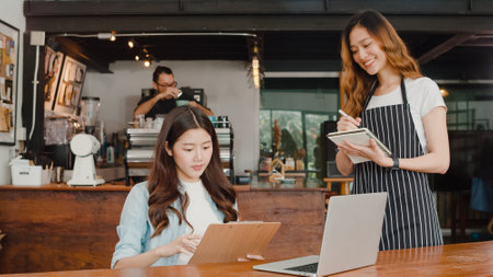 Cheerful Asian lady waitress with notebook taking receive order menu from young client girl at urban cafe. Young Asian freelance women working on laptop at coffee shop. Owner small business concept.の写真素材
