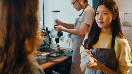 Young Asia female barista waiter taking order from customer standing behind bar counter while talking with customer making note on takeaway coffee cup at cafe restaurant. Owner small business concept.の写真素材