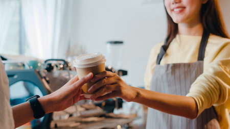 Young Asia female barista serving take away hot coffee paper cup to consumer standing behind bar counter at cafe restaurant. Owner small business, food and drink, service mind concept.の写真素材
