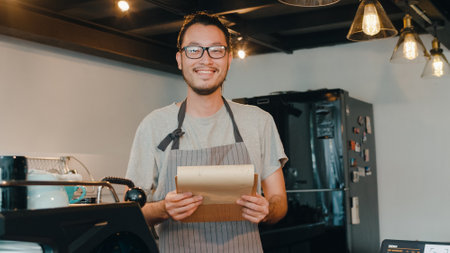 Portrait young Asian guy barista take receive order with menu feeling happy at urban cafe. Asia small business owner boy in apron relax toothy smile looking to camera stand at counter in coffee shop.の写真素材