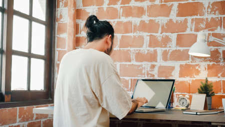 Back view freelance hipster modern Asian man designer working home using laptop in living room at home. Happy young male work while sitting at desk in creative office. Enjoying time at home concept.の写真素材