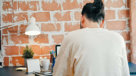 Back view freelance hipster modern Asian man designer working home using laptop in living room at home. Happy young male work while sitting at desk in creative office. Enjoying time at home concept.の写真素材