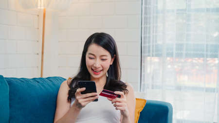Young smiling Asian woman using smartphone buying online shopping by credit card while lying on sofa when relax in living room at home. Lifestyle women at house concept.の写真素材