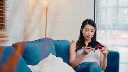 Young smiling Asian woman using smartphone buying online shopping by credit card while lying on sofa when relax in living room at home. Lifestyle women at house concept.の写真素材