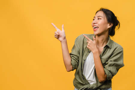Portrait of young Asian lady smiling with cheerful expression, shows something amazing at blank space in casual clothing and standing isolated over yellow background. Facial expression concept.の写真素材