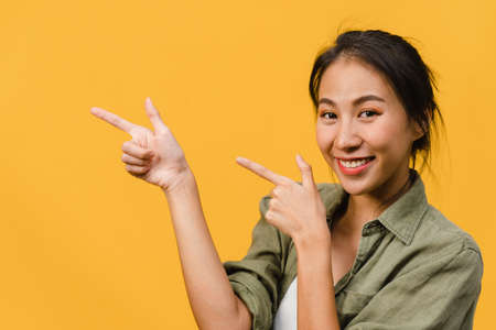 Portrait of young Asian lady smiling with cheerful expression, shows something amazing at blank space in casual cloth and looking at camera isolated over yellow background. Facial expression concept.の写真素材