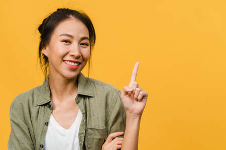Portrait of young Asian lady smiling with cheerful expression, shows something amazing at blank space in casual cloth and looking at camera isolated over yellow background. Facial expression concept.の写真素材