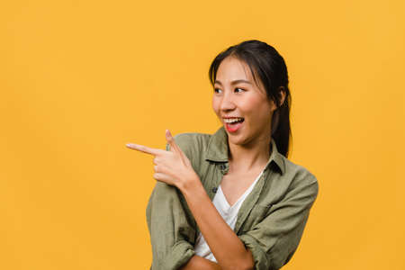Portrait of young Asian lady smiling with cheerful expression, shows something amazing at blank space in casual clothing and standing isolated over yellow background. Facial expression concept.の写真素材