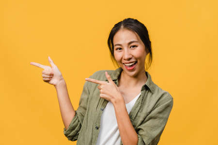 Portrait of young Asian lady smiling with cheerful expression, shows something amazing at blank space in casual cloth and looking at camera isolated over yellow background. Facial expression concept.の写真素材