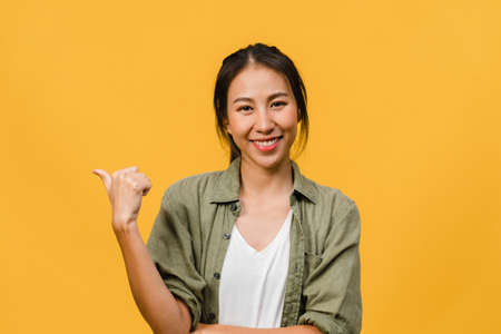 Portrait of young Asian lady smiling with cheerful expression, shows something amazing at blank space in casual cloth and looking at camera isolated over yellow background. Facial expression concept.の写真素材