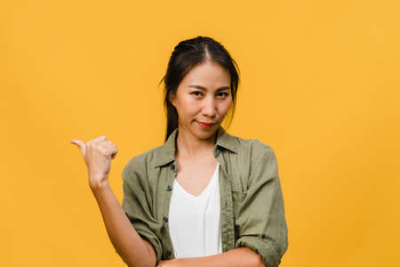 Portrait of young Asian lady smiling with cheerful expression, shows something amazing at blank space in casual cloth and looking at camera isolated over yellow background. Facial expression concept.の写真素材