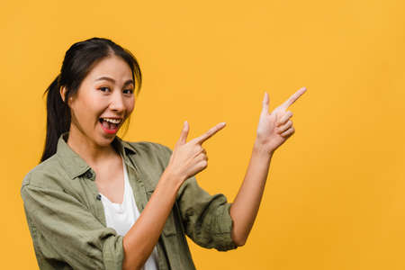 Portrait of young Asian lady smiling with cheerful expression, shows something amazing at blank space in casual cloth and looking at camera isolated over yellow background. Facial expression concept.の写真素材