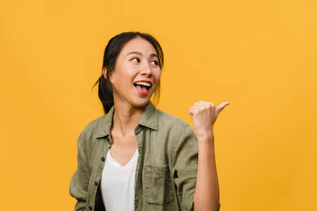 Portrait of young Asian lady smiling with cheerful expression, shows something amazing at blank space in casual clothing and standing isolated over yellow background. Facial expression concept.の写真素材