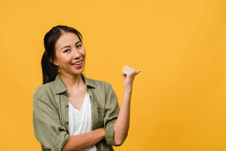 Portrait of young Asian lady smiling with cheerful expression, shows something amazing at blank space in casual cloth and looking at camera isolated over yellow background. Facial expression concept.の写真素材