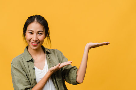 Portrait of young Asian lady smiling with cheerful expression, shows something amazing at blank space in casual cloth and looking at camera isolated over yellow background. Facial expression concept.の写真素材