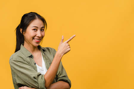 Portrait of young Asian lady smiling with cheerful expression, shows something amazing at blank space in casual cloth and looking at camera isolated over yellow background. Facial expression concept.の写真素材