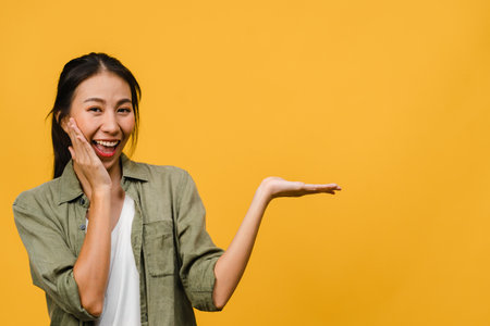 Portrait of young Asian lady smiling with cheerful expression, shows something amazing at blank space in casual cloth and looking at camera isolated over yellow background. Facial expression concept.の写真素材