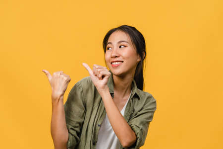Portrait of young Asian lady smiling with cheerful expression, shows something amazing at blank space in casual clothing and standing isolated over yellow background. Facial expression concept.の写真素材
