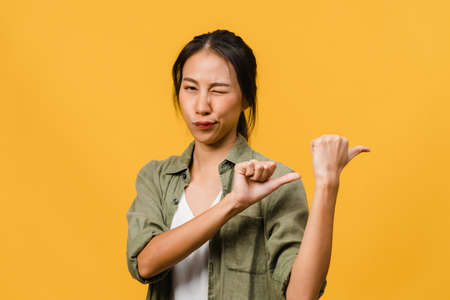 Portrait of young Asian lady smiling with cheerful expression, shows something amazing at blank space in casual cloth and looking at camera isolated over yellow background. Facial expression concept.の写真素材
