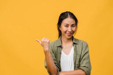Portrait of young Asian lady smiling with cheerful expression, shows something amazing at blank space in casual cloth and looking at camera isolated over yellow background. Facial expression concept.の写真素材