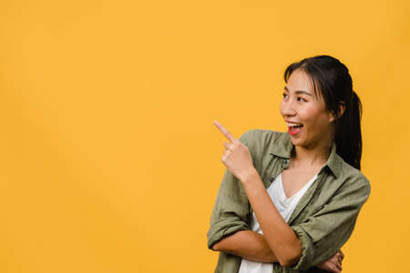 Portrait of young Asian lady smiling with cheerful expression, shows something amazing at blank space in casual clothing and standing isolated over yellow background. Facial expression concept.の写真素材