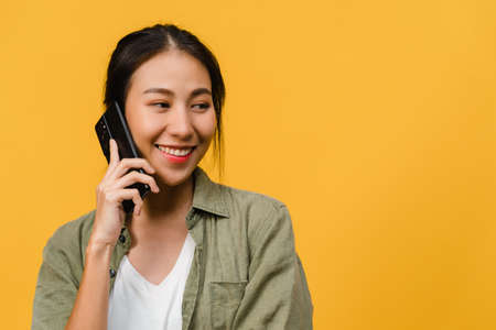 Young Asia lady talk by phone with positive expression, smile broadly, dressed in casual clothing feeling happiness and stand isolated on yellow background. Happy adorable glad woman rejoices success.の写真素材