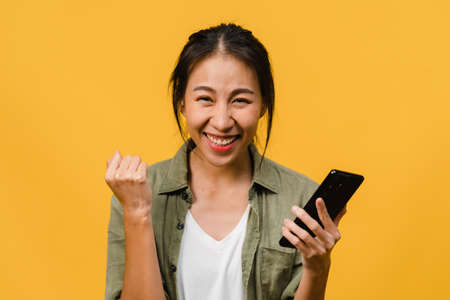 Surprised young Asia lady using mobile phone with positive expression, smiles broadly, dressed in casual clothing and stand isolated on yellow background. Happy adorable glad woman rejoices success.の写真素材