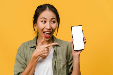 Young Asia lady show empty smartphone screen with positive expression, smiles broadly, dressed in casual clothing feeling happiness on yellow background. Mobile phone with white screen in female hand.の写真素材