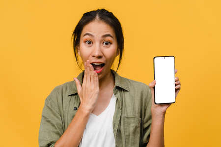 Young Asia lady show empty smartphone screen with positive expression, smiles broadly, dressed in casual clothing feeling happiness on yellow background. Mobile phone with white screen in female hand.の写真素材