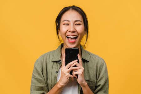 Surprised young Asia lady using mobile phone with positive expression, smile broadly, dressed in casual clothing and looking at camera on yellow background. Happy adorable glad woman rejoices success.の写真素材