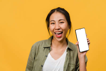 Young Asia lady show empty smartphone screen with positive expression, smiles broadly, dressed in casual clothing feeling happiness on yellow background. Mobile phone with white screen in female hand.の写真素材