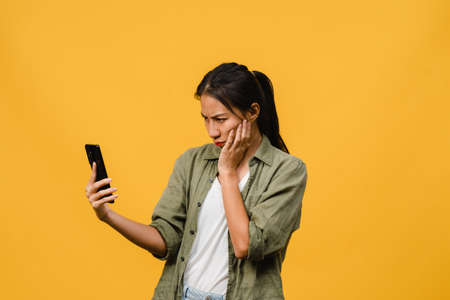 Thinking dreaming young Asia lady using phone with positive expression, dressed in casual cloth feeling happiness and stand isolated on yellow background. Happy adorable glad woman rejoices success.の写真素材