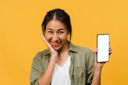 Young Asia lady show empty smartphone screen with positive expression, smiles broadly, dressed in casual clothing feeling happiness on yellow background. Mobile phone with white screen in female hand.の写真素材