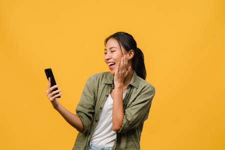 Young Asia lady using phone with positive expression, smiles broadly, dressed in casual clothing feeling happiness and stand isolated on yellow background. Happy adorable glad woman rejoices success.の写真素材